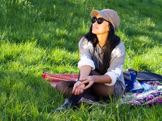 Beautiful Asian woman wearing a hat and sunglasses having a relaxing time in the city park sitting on a colorful blanket surrounded with a green grass.