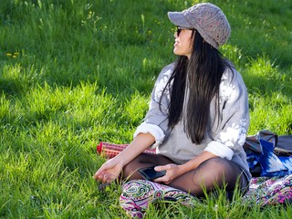 Beautiful Asian woman wearing a hat and sunglasses having a relaxing time in the city park sitting on a colorful blanket surrounded with a green grass. 