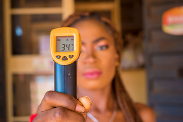 Close-up shot of a young african woman using infrared forehead thermometer (thermometer gun) to check her body temperature for virus symptoms - epidemic virus outbreak concept