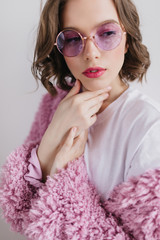 Amazing girl with pink lips posing in fur coat in studio. Portrait of lovely young woman with wavy hair standing on white background.