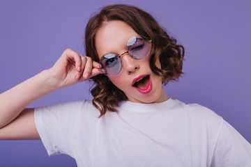 Cheerful white girl in sunglasses playfully posing on bright purple background. Indoor photo of sensual curly young woman looking to camera.
