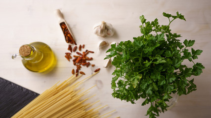 Top dpwn flat lay view of the typical italian recipe spaghetti aglio olio e peperoncino (garlic, oil and hot pepper) ingredients with a glass jar with sprigs of parsley on light wooden table