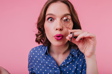 Close-up shot of brown-eyed lady with surprised face expression curls her eyelashes. Studio portrait of lovable caucasian girl with short hair preparing for date.