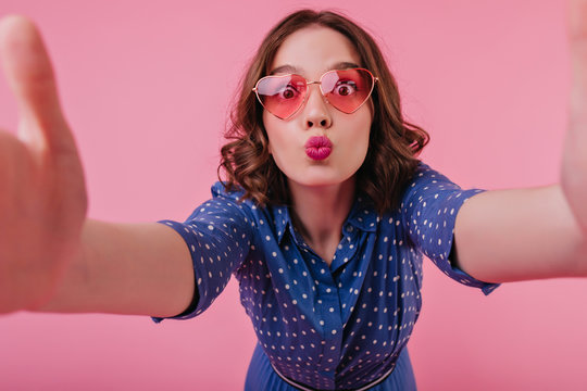 Positive surprised girl with tattooes making selfie with kissing face expression. Adorable brunette woman in blue blouse and glasses taking picture of herself.