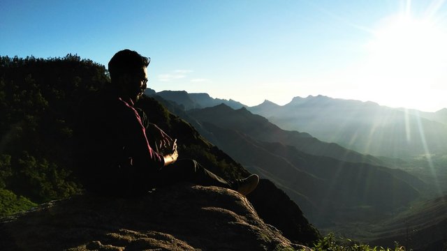 Rear View Of Male Hiker Sitting On Mountain Against Sky During Sunny Day