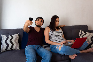 Young adult couple sitting on sofa, husband watching tv and wife working from home on laptop seriously