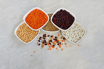 A set of different dried legumes in ceramic bowls: red lentils, green lentils, peas, red beans, white beans on grey background