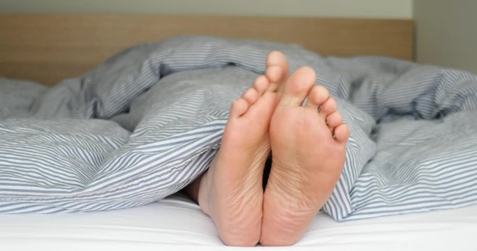 Pair of Caucasian male feet sole sticking out of the quilt in bed relaxing and rubbing against each other