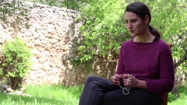 Concentrated young woman praying the rosary on a chair in the garden on a sunny day.