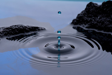 Gotas de agua color azul congeladas en el aire y formando ondas en la superficie liquida, con rocas...