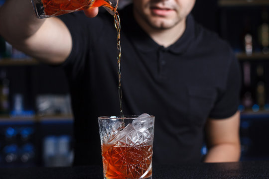 Cocktail negroni americano poured by barmen into vintage elegant rock glass old fashioned whiskey glass. Popular alcoholic drink made of vermouth and gin. Bartender in black top in background.