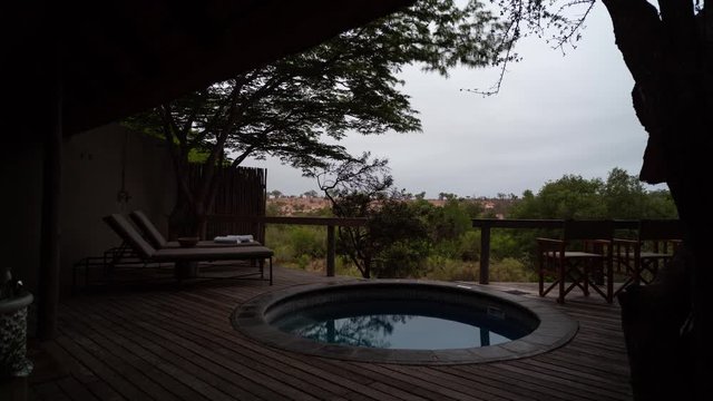 Static early morning timelapse on cloudy overcast morning, view from lodge room splash pool in nature, deck chairs and coffee as the day breaks in bushveld/reserve, framed with Brack thorn tree.