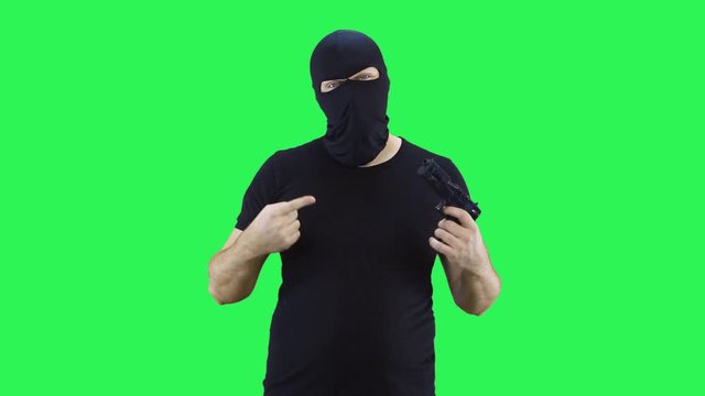A masked man speaks to the camera, threatens with a gun.Balaclava.Green screen background.