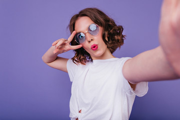Carefree young lady with wavy hairstyle making selfie on bright purple background. Indoor shot of lovely european girl with dark hair posing in glasses with kissing face expression.