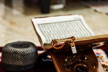 Quran, the Scripture of Muslims on rihal or wood book stand, prayer beads or tasbeeh, skullcap hat, at home.  The concept of staying home in ramadan during a corona virus disease pandemic situation.