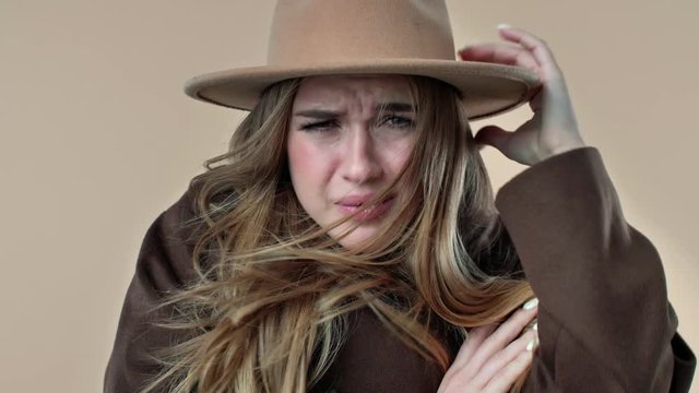 A close-up view of a displeased young woman wearing hat and coat is feeling cold isolated over the beige background in studio