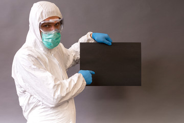 Doctor with surgical mask, goggles and protective suit pointing a black banner on gray backround.