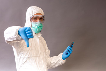 Male doctor with surgical mask, goggles and protective suit holding his phone and pointing okay on gray background.