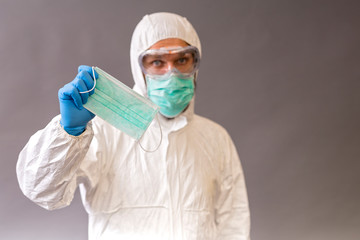 Male doctor with surgical mask, goggles and protective suit holding a medical mask on gray background.