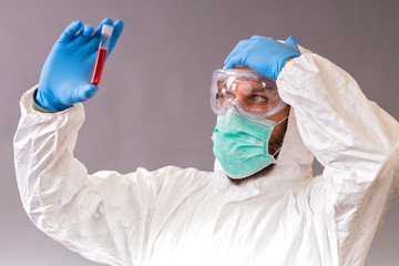 Male doctor with surgical mask, goggles and protective suit holding blood samples on gray backround. Vials with cure Covid-19.