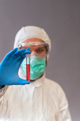 Male doctor with surgical mask, goggles and protective suit holding blood samples on gray backround. Vials with cure Covid-19.