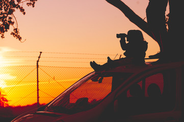 Silhouette against a sunset. Man with children on car roof observing something in the distance through binoculars.