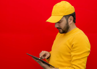 Home delivery worker in yellow hat, blank t-shirt, holding tablet, isolated on red background. Courier service worker.