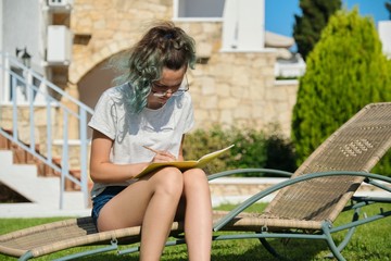 Girl teenager 15, 16 years old sitting on lounger on lawn, writing with pencil in notebook