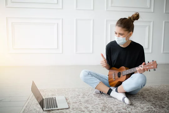A boy wearing a face mask plays ukulele in front of a laptop Stock