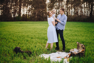 Family in nature. Picnic in the forest, in the meadow. Green grass. Blue clothes. Mom, dad, son with glasses. Boy with blond hair. Joy. Parents play with child. Together. Picnic basket. Food, blanket