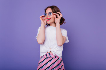 Happy cute girl with wavy hairstyle touching her glasses with smile. Indoor shot of magnificent curly lady in white t-shirt posing on purple background.