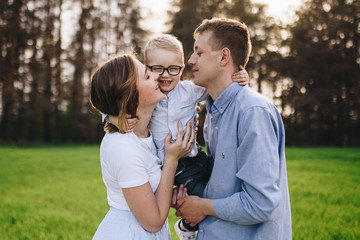 Fototapeta premium Family in nature. Picnic in the forest, in the meadow. Green grass. Blue clothes. Mom, dad, son with glasses. Boy with blond hair. Joy. Parents play with child. Together. Picnic basket. Food, blanket