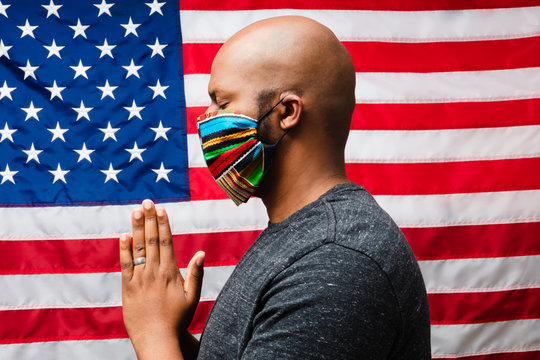 Black man wearing cloth mask and praying in front of American flag