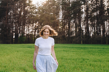 Fototapeta na wymiar The girl is blonde, brown hair, in a white shirt and blue midi skirt. Walking in the field, through the green grass. Portrait of a girl. Positive and smile.