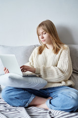 Cute young female manager working via laptop, hipster girl in white sweater and jeans sitting on a bed using modern notebook. Concept of online work, studying or freelance during quarantine