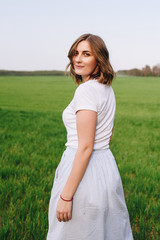 Fototapeta na wymiar The girl is blonde, brown hair, in a white shirt and blue midi skirt. Walking in the field, through the green grass. Portrait of a girl. Positive and smile.