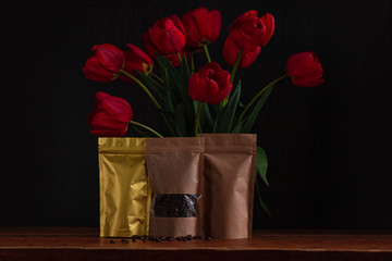 Pouch bags for coffee and tea with amazing blooming red tulips bouquet on black background. Food shop packaging mockup. Foiled metal and kraft paper packs. Dark moody low key flowers banner copy space