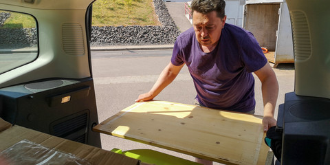 Plump,dark-haired,middle-aged man unloads wooden planks from trunk of compact MPV to dump it at station of collection of large-size garbage in Germany,Europe.Spring sunny day.Selective focus,close up