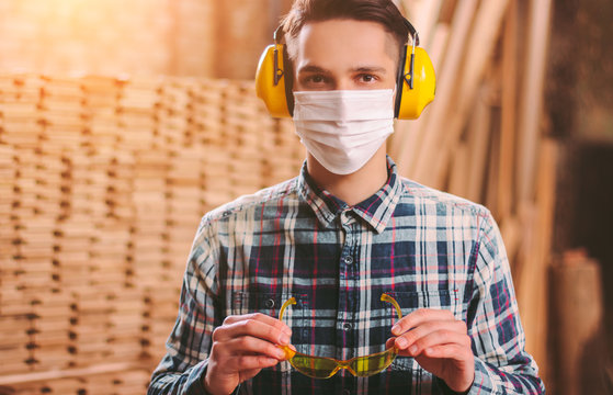 Portrait of young carpenter in hearing protectors headphones and medical face mask wearing protective eyewear at workshop. Male craftsman at sawmill. Factory work at coronavirus COVID-19 quarantine