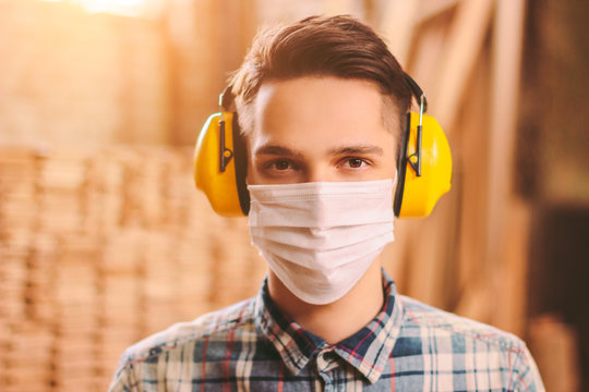 Portrait of confident man carpenter in medical face mask and hearing protectors at wooden workshop. Professional craftsman in protective mask and headphones at sawmill. Young male artisan at workplace - Powered by Adobe