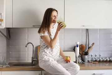 eautiful girl drinks freshly prepared smoothie in kitchen. smoothies freshly made from assorted vegetable ingredients on kitchen counter. Healthy Eating. selective focus