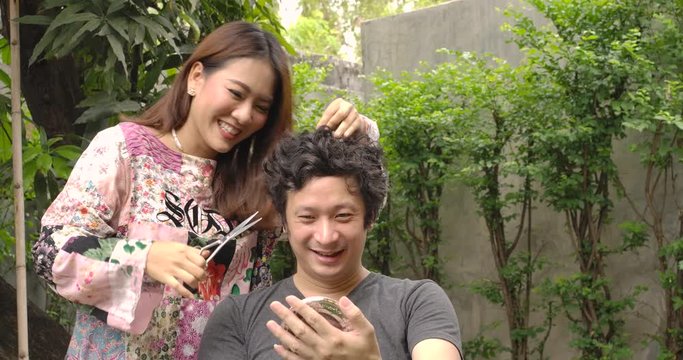 Young Asian Woman Trying To Does Haircuts To Her Boyfriend Or Husband With Scissors At Home Because Of Lockdown. Stay At Home Quarantine Coronavirus Pandemic Prevention, Social Isolation Concept.