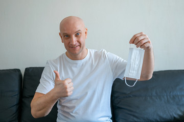 Portrait of a bald man sitting at home on the sofa holding a medical mask in his hands