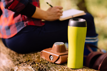 stylish hipster young woman sitting on a tree trunk, with a backpack, thermo mug and notebook in the forest on sunset light in the spring season, looking at amazing woods, travel concept