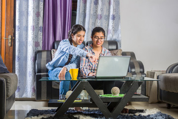 two girls sitting in a living room using  laptop and books for online classes