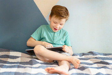 Fototapeta premium a cheerful child sits on a bed with a plaid blanket, runs his finger over the tablet screen and smiles