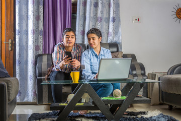 two girls sitting in a living room using  laptop and books for online classes