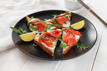 Sandwich with salmon, cheese, arugula and lemon on a black plate on a white table