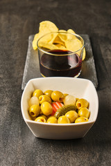 Closeup of olives with pickles, mini onions, carrot and chips in a white bowl, wine in a crystal glass on black slate. Brown wood background. Home or bar appetizer.