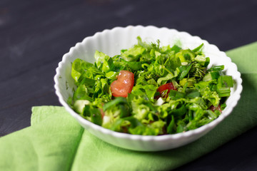 salad with arugula, cucumber, greenery and tomatoes in white bowl on green textile on black wooden background